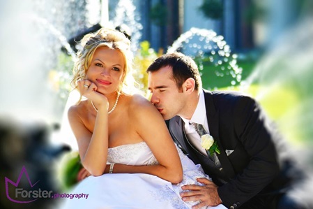 Ein Brautpaar im Hochzeits-Outfit sitzt beim Fotoshooting vor einem Brunnen, er küsst sie zärtlich auf die Schulter