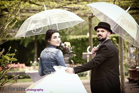 Ein Brautpaar im Hochzeits-Outfit steht beim Fotoshooting im Regen in einem Park und tragen zwei Regenschirme