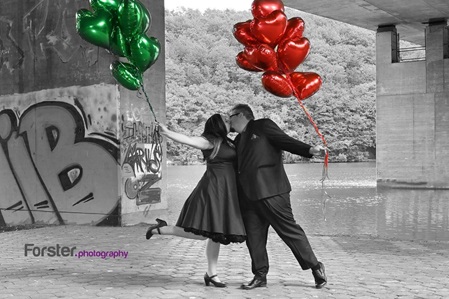 Ein elegantes Brautpaar küsst sich beim Fotoshooting mit roten und grünen Luftballons am See unter einer Brücke