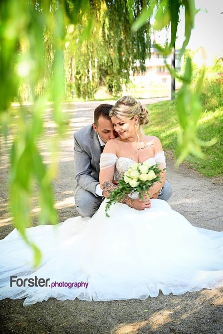 Eine Braut sitzt mit Bräutigam beim Fotoshooting alleine mit Brautstrauß auf einer Wiese. Das weiße Kleid ist ausgebreitet